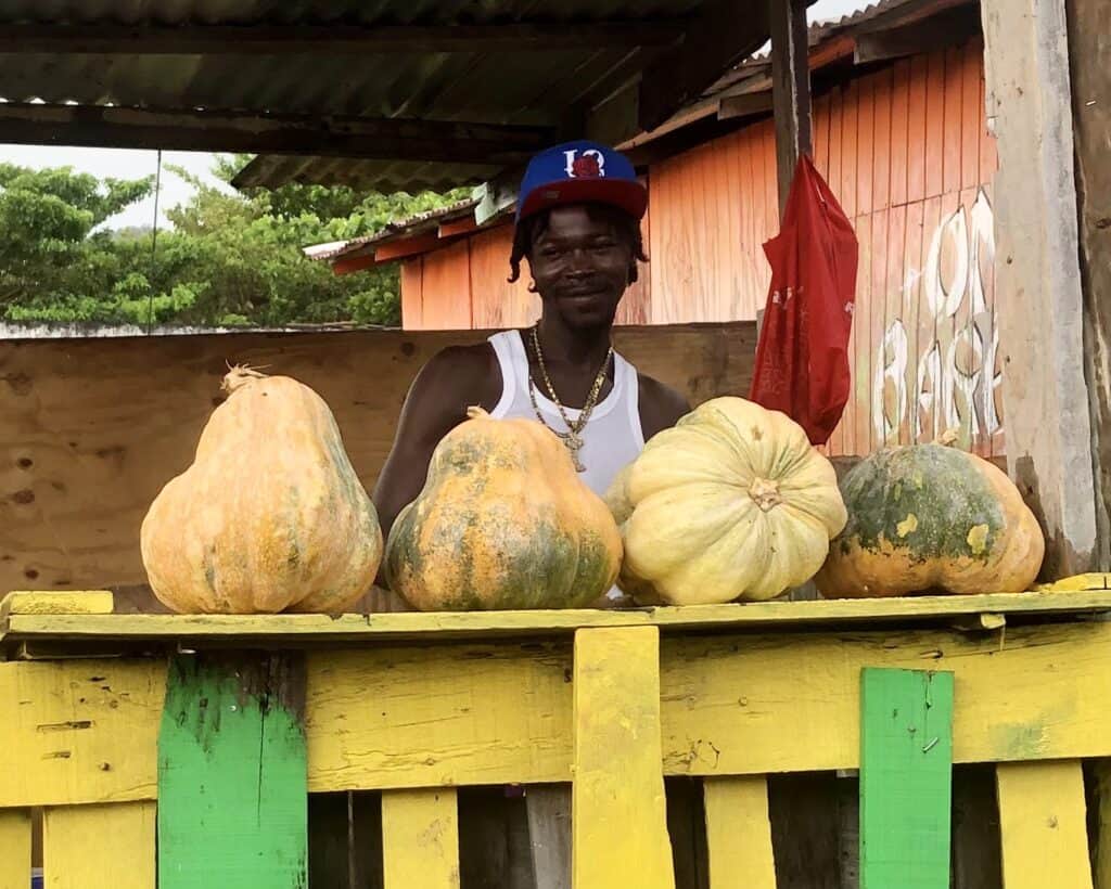 Pumpkin Man smiling with a stall of bright orange pumpkins outside Sauteurs market.